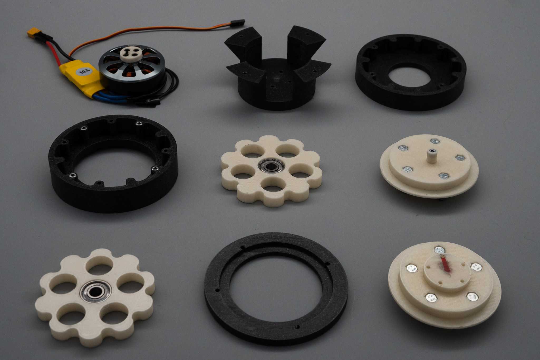 All the parts of the gearbox laid out on a gray surface: a brushless motor with a yellow 30 amp speed controller, black housing pieces, two cream cycloidal discs with center bearings, and two cream output plates with hex bolts.