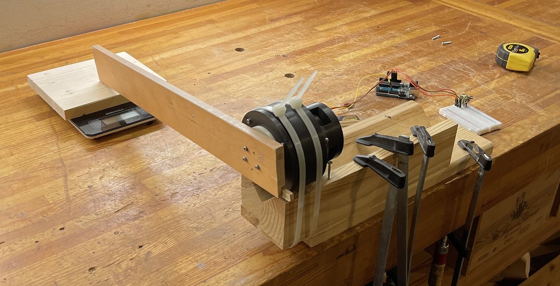 A bench torque-test rig: the black gearbox zip-tied into a wooden cradle clamped to a workbench, a long wooden lever arm on its output resting on a digital scale, with an Arduino, a breadboard, and a tape measure alongside.